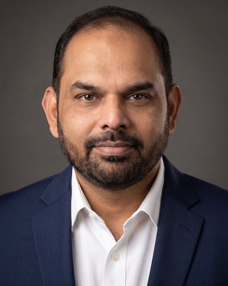Studio portrait of Dr Suraj Menon in a dark suit and white shirt