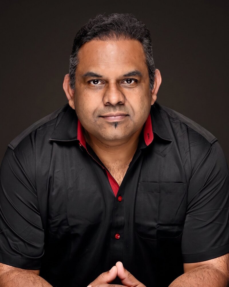 Studio portrait of Dr Menon in a black shirt with red trim, arms crossed